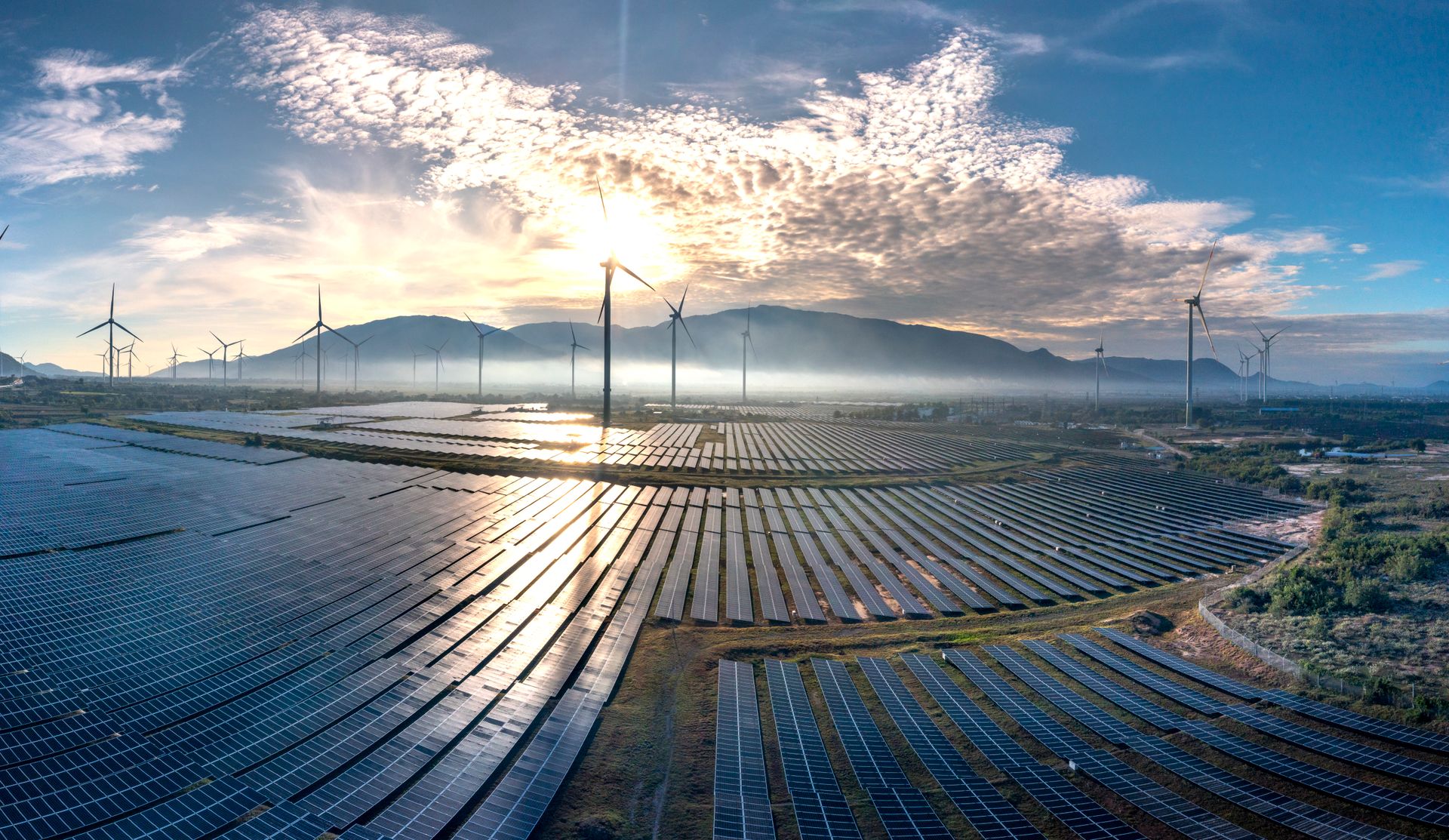 Vista aérea de uma usina solar fotovoltaica com turbinas eólicas ao fundo, sob a luz dourada do amanhecer.